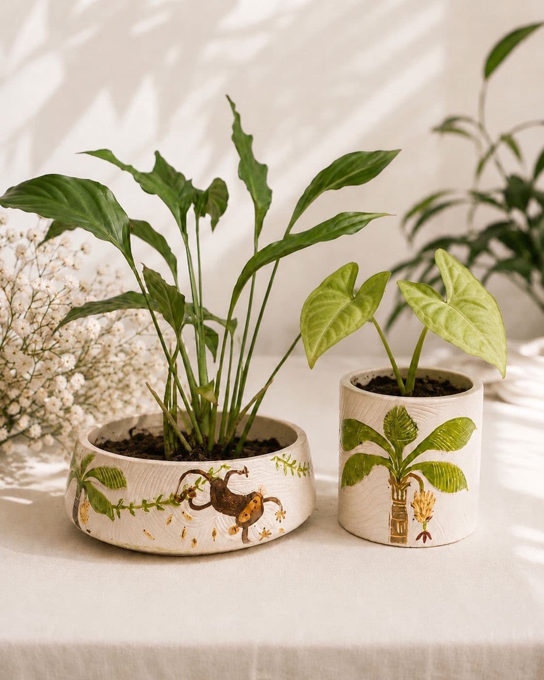 A wide carved bowl planter and a taller cylinder pot, painted with a monkey and a banana palm, planted up on a linen tablecloth in afternoon light